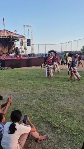 The video shows an outdoor performance featuring several dancers in traditional costumes. The dancers wear patterned shirts, white skirts, and elaborate headpieces resembling horses. They are moving in unison on a grassy field. In the background, there is a stage with lighting equipment and speakers, suggesting a performance setting. Spectators are visible in the foreground, with one person sitting and recording the event on a phone. The sky is clear and blue, indicating daylight.