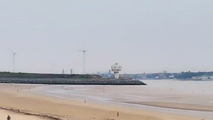 The video shows a beach with sand dunes. In the distance, there is a breakwater with a lighthouse and some wind turbines. Beyond that, a port with several large cranes is visible, along with some buildings. The sky is overcast. There are a few people walking on the beach.