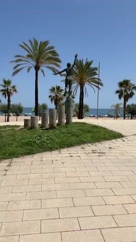 A bronze statue of a young man in shorts with his right arm raised and left arm extended stands on a pedestal. The statue is positioned on a grassy area with several concrete cylindrical posts of varying heights arranged in front of it. In the background, there are palm trees, a beach with people, and the ocean under a clear blue sky. The foreground is paved with light-colored bricks.