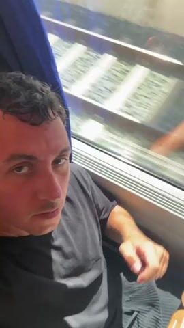 A person is filming themselves from the chest up while sitting on a train, looking towards the camera with a neutral expression. They are wearing a black t-shirt and have short, dark, curly hair. The person raises their right hand towards the camera, making a fist. In the background, through a window, train tracks can be seen moving rapidly, indicating the train is in motion. The person's left hand rests on the edge of a table, where a tray of food is visible. The camera then pans to a close-up of the person's face as they smile, and then shifts to show the interior of the train carriage, with other passengers seated. The video continues with the person in the foreground, now wearing a blue t-shirt, turning their head away from the camera. The video ends with a blurry close-up of the person's face.