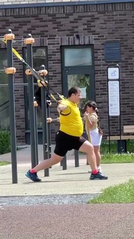 A man in a yellow t-shirt and black shorts is performing an exercise with yellow resistance straps attached to a black metal structure. He is in a lunge position, pulling the straps towards his chest. A child wearing a light-colored t-shirt and white shorts stands behind him, also holding onto a resistance strap, facing away from the camera. They are outdoors on a paved area with grass and a brick building in the background.
