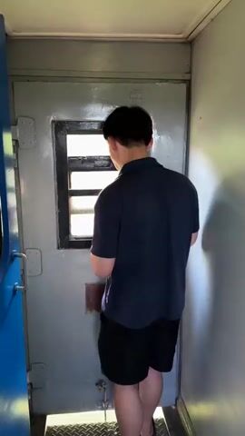 A person wearing a dark blue polo shirt and black shorts stands facing a window in a train car. The person is looking out the window and appears to be holding something in their hands near their face. The train car interior is visible with a gray door and white walls. The window has bars. Sunlight is coming through the window.