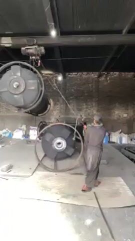 In an indoor industrial setting, a person clad in gray clothing and a dark cap is shown operating an overhead hoist. The hoist suspends a large, dark object from the ceiling. The object appears to be a cylindrical industrial component with a circular fan-like structure at one end. The device is being lowered towards the ground, where a similar object is already resting. The environment is characterized by a corrugated metal ceiling, brick walls, and a concrete floor. There is equipment and miscellaneous materials located in the background, further emphasizing the industrial nature of the scene.