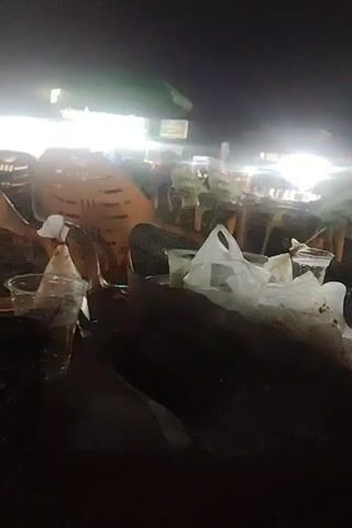 The video shows a night scene at what appears to be an outdoor food stall area. There are multiple orange plastic chairs, some stacked, and tables with plastic cups, plastic bags, and food remnants on them. The area is illuminated by bright, white lights from the stalls. The camera is mostly static, providing a view from a low angle, focusing on the nearby table. The overall scene suggests a casual, outdoor dining or gathering spot.