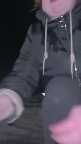 A young woman interacts with a large dog in an outdoor environment at night. The woman, dressed in dark clothing, kneels down and pets the dog. The dog, primarily brown with darker shading, has glowing eyes in the dark. The ground appears to be covered with snow or a light-colored substance. The scene is dark with only a hint of background visibility.