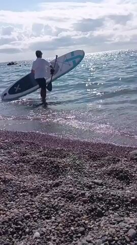 A person, wearing a white shirt and dark shorts, walks into the water carrying a paddleboard. The person then kneels on the paddleboard and begins to paddle away from the shore. The water is clear and the sky is partly cloudy. The shore is covered in small stones.