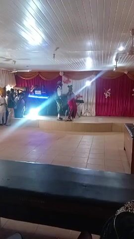 The video shows a man and a woman on a stage. The man is wearing a polka-dotted vest, star-printed pants, and a headscarf. The woman is wearing a long red dress. They appear to be dancing together. Behind them, there's a DJ setup with blue lights, and a backdrop consisting of gold and red drapes. The stage is tiled. The room has a low, white ceiling with ceiling fans. Other people are seen standing near the DJ setup.