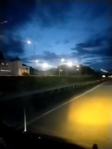 The video shows a view from a vehicle driving on a highway at dusk or night. The sky is dark blue with some clouds, and streetlights illuminate the road. There are streetlights on the side of the highway, and some appear to be on poles. A hill or mountain is visible in the background. The headlights of the vehicle are on, casting a yellow glow on the road. There is a building with arches visible on the left side of the frame. The road has white lane markings.