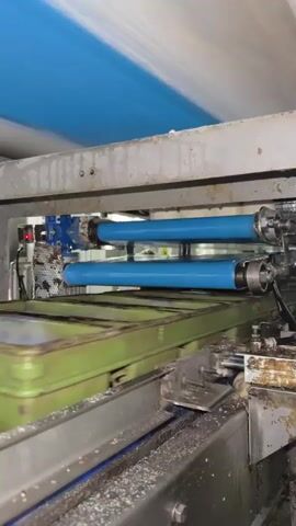 The video shows an industrial food processing machine in operation. Two blue rollers are rotating, with what appears to be dough or a similar food product being fed between them. The product is blurry due to its rapid movement. The machine has a metal frame, and there is a green tray beneath the rollers containing more of the food product. White debris is visible on the conveyor belt below. The lighting suggests an indoor, factory environment. The overall impression is of an automated process for shaping or processing food items.