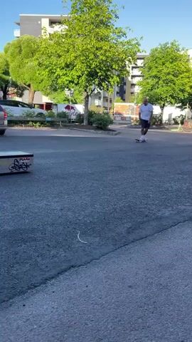 This video captures a skateboarder performing tricks in an outdoor urban setting, likely in Karlstad, Sweden. The scene appears to be a parking lot or an area adjacent to a residential or commercial building. The time of day is late afternoon or early evening, indicated by the long shadows and the clear blue sky with scattered clouds. The weather seems pleasant, with the sun shining and trees in full leaf.

The primary subject is a man in a white t-shirt and dark shorts, wearing white socks and sneakers, actively skateboarding. He is captured mid-trick, leaping over a low obstacle, which appears to be a skate ramp or a similar structure. This ramp has graffiti on its side, including the word "PLAZA" repeated several times, along with other artistic tags.

In the background, several cars are parked, including a silver station wagon and a black sedan. Behind the cars and trees, modern apartment buildings with balconies are visible, suggesting a residential area. The overall mood is one of casual recreation and urban sports. The focus is on the skateboarder's movement and skill, with the environment serving as a backdrop to his activity. No specific dialogue or audio is present beyond the ambient sounds of the environment. The presence of a street sign with a blue pedestrian symbol also adds to the urban context.