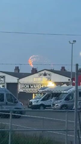 The video shows a building with the sign "IRON WAREHOUSE" across its front. Above the sign, there is a logo depicting a barbell with weights. On the roof of the building, a display of fireworks is visible, with sparks and streaks of light against a darkening sky. Several vehicles, including vans and what appear to be campers or RVs, are parked in front of the building. A chain-link fence is in the foreground, partially obscuring the view. The overall atmosphere suggests an outdoor scene at dusk or early evening.