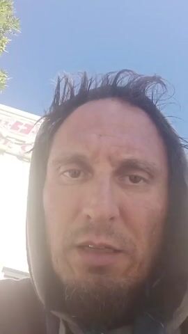 A man is speaking directly to the camera. He has dark hair and a goatee. He is wearing a grey hoodie. The sky is blue. A building or structure is visible in the background.