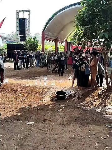 The video shows an outdoor event with a group of people gathered. A tent with red and white fabric is set up in the background, and large speakers are visible to the left. The ground is muddy and appears wet. Several children are in the foreground, some being splashed with water. A black bucket is on the ground. Adults are standing around, observing. Some individuals in the center appear to be engaged in an activity that involves splashing water. The overall atmosphere seems to be a communal gathering with some form of water play or ceremony.