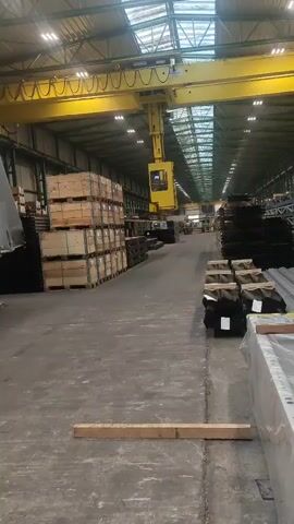This video is a first-person perspective, likely from a smartphone, looking down a long, industrial warehouse. The camera is slightly angled upwards, showing a large yellow overhead crane with a yellow operator cab suspended beneath it. The crane is stationary at the moment, but the hook assembly is visible. To the left of the frame, there are stacks of wooden crates. To the right, there are stacks of what appear to be metal goods or lumber. The floor is concrete, and there are wooden planks lying on the ground in the foreground. The warehouse has a high ceiling with multiple skylights or translucent panels that allow natural light to enter, supplemented by rows of artificial lights. The overall impression is of an organized industrial storage or manufacturing facility. The video is short, showing no discernible action or movement beyond a slight camera shake.