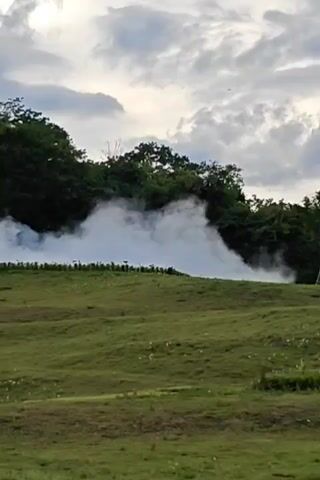 The video shows a hillside covered in green grass. At the top of the hill, a line of dark green trees is visible. A large plume of white smoke or mist is billowing from behind the trees, obscuring some of the vegetation. The sky above is partly cloudy with a mix of white and grey clouds. The overall impression is of a natural landscape with an unusual atmospheric phenomenon occurring.