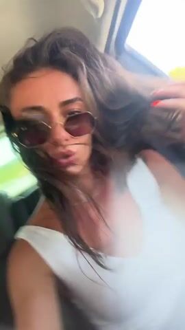 A woman with long, dark hair and sunglasses is in the passenger seat of a car. Her head is tilted back, and she appears to be laughing. The car is moving, and trees and sky are visible through the open sunroof.