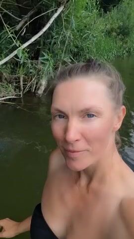 A woman with blonde hair pulled back in a bun and blue eyes smiles at the camera while in the water. She is wearing a black swimsuit top. The water is murky green, and the background shows lush green trees and vegetation along the bank of a body of water. The video appears to be shot from a selfie perspective, with the camera submerged in the water. The woman's face is partially obscured by the water, with ripples and bubbles visible. The scene suggests a natural outdoor environment, possibly a river or lake.
