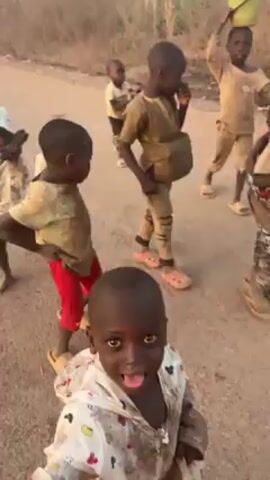 The video shows a group of young African children on a dirt road, possibly in a rural area. They are mostly wearing dirty clothes and some have items on their heads or in their hands, like containers. The children are smiling and looking at the camera. In the background, there are trees and possibly a power line.