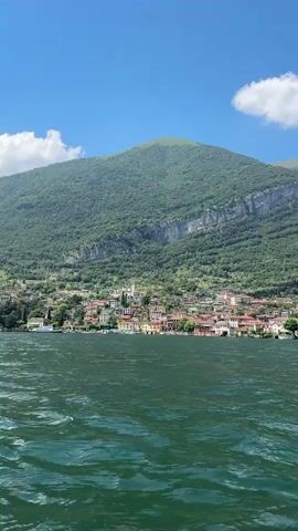The video shows a wide shot of a scenic lake with a small village nestled at the base of a lush green mountain. The water in the foreground is a deep green and appears to be choppy, with visible waves. In the middle ground, a line of colorful buildings forms the village, with a church steeple visible among them. The village is situated on the shore of the lake, and a few boats are visible in the water near the village. Behind the village, a large, densely forested mountain rises steeply into a clear blue sky with a few white clouds. The overall impression is of a tranquil and picturesque natural setting with a charming human settlement.