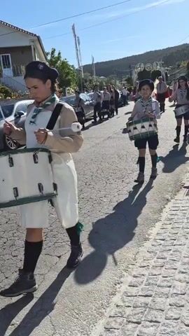 The video shows a procession marching down a cobblestone street on a sunny day. The group consists of uniformed scouts, most of whom are playing drums. The scouts are wearing khaki shirts, dark skirts or shorts, berets, and knee socks.  The drums are strapped across their chests, and they are using drumsticks to play a steady beat. Behind the scouts are other individuals, presumably adults, walking in the procession. There are cars parked along the side of the street, and houses are visible in the background. The road shows signs of wear and cracks. The shadows of the marchers are long and distinct on the street due to the bright sunlight. The overall atmosphere is of a ceremonial or celebratory march.
