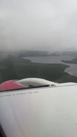 The media file depicts an aerial view from the window of an airplane, likely during takeoff or landing. The scene outside the window reveals a vast expanse of dark green forest interspersed with numerous lakes and bodies of water. The landscape is enveloped in a thick, overcast sky, suggesting cloudy or foggy weather conditions.

In the foreground, the wing of the airplane is visible, along with a portion of the engine nacelle, which is painted pink and white. There is some text visible on the wing, written in Russian. It reads: "НЕ ВСТУПАЙ ЗА ПРЕДЕЛЫ ЭТОЙ ЗОНЫ" which translates to "DO NOT STEP BEYOND THIS ZONE."

The overall atmosphere is subdued, due to the cloudy weather and the muted colors of the landscape. The time of day is unclear, but the overcast conditions might suggest a diffused light. The location context points to Murmashi, Russia, which aligns with the forested and lake-filled terrain. There are no people or specific objects other than the airplane itself and the natural scenery. The activity taking place is simply the flight of the airplane over this specific geographical area.