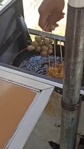 A hand is seen holding several skewers of food being fried in a pan of oil. The skewers contain round, brownish food items and some pieces that appear to be breaded and golden brown. The pan is set in a cooking cart with a metal pole visible in the foreground. The background is blurred but suggests an outdoor setting with sunlight.