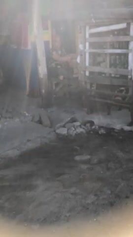 A shaky video shows a makeshift stall with wooden shelves. A person wearing a red hat and a blue garment is visible on the left. Another person wearing camouflage clothing is standing near the stall, holding something to their mouth. The ground appears to be dirt and rocks. The stall has a tarp overhead, and shelves are stocked with various items, though they are blurry. The overall scene suggests an outdoor market or informal shop.