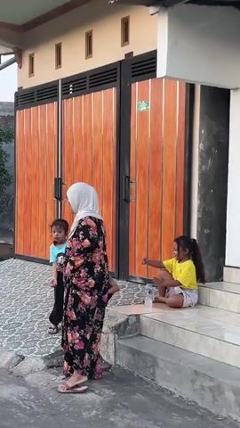 A woman wearing a hijab and a floral dress stands on a patterned concrete surface in front of a large wooden gate with black accents. To her left, a young child in a blue shirt and dark pants stands. To her right, on a set of white stairs leading up to a building, a young girl in a yellow shirt and shorts sits, holding a bottle of water and gesturing with her right hand. The gate is closed. The environment appears to be outdoors, possibly a residential area.
