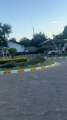 The video shows a view of the front of Sunbird Academy. The scene is outdoors, with a paved driveway and well-maintained landscaping featuring a flowerbed at the end of the drive. A man can be seen walking towards the building in the background. There are some parked cars nearby. The sky is blue with some clouds. The video is taken from a static position, and there are no audio elements to analyze.