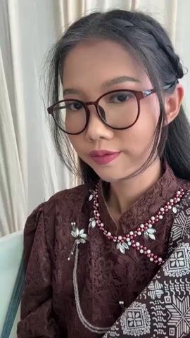 A medium shot captures a woman sitting indoors, looking into the camera. She wears glasses, a maroon lace top, and a patterned scarf, adorned with a silver brooch and a necklace. Her dark hair is styled with a braid and loose strands framing her face. The background features light-colored curtains and an unseen speaker's voice. The woman maintains eye contact, then slightly bows her head, glancing down and away from the camera.