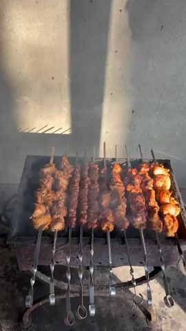 The video shows several skewers of meat cooking over coals in a grill. The meat is arranged on metal skewers and appears to be shish kebab. Smoke is rising from the grill, indicating the meat is actively cooking. The background consists of a concrete wall, and there is sunlight casting shadows on the wall and the grill. The overall scene depicts an outdoor cooking event.