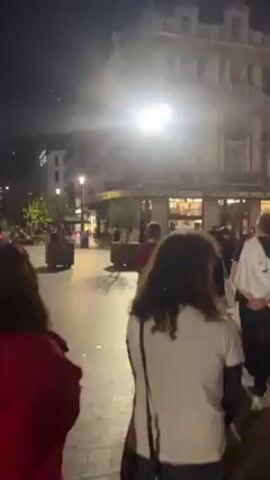 The video depicts a bustling city square at night, illuminated by streetlights and building lights. Numerous people are seen walking around and congregating. The background features a grand building with large columns and banners hanging from its facade. Several restaurants and bars appear to be open, with patrons visible inside. A bicycle lies on the ground.