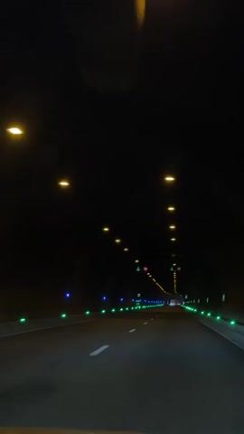 The video shows the perspective from inside a car driving through a dark tunnel. The tunnel is lit by yellow overhead lights and green and blue lights along the sides of the road. There are white lines marking the lanes on the road. There are speed limit signs visible, one with a 70 inside a red circle and another with a 50 inside a red circle, both with green arrows pointing downwards. The tunnel appears to be long, stretching far into the distance. The only sounds are some loud electronic music.