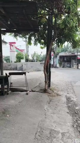 The video begins with a view of a dirt path and a wooden shelter with a table and chairs underneath. A tree stands to the right of the shelter. In the background, there is a building with a red and white facade. The camera then pans down to show a person's legs and feet wearing sandals and black pants with a blue stripe. The camera then moves forward, showing a motorcycle's handlebar and mirror. The scene shifts to a street with a small food stall. A woman in a headscarf is standing at the stall, which has a sign that reads "PAFFTA FRIED CHICKEN". Another person is on a motorcycle to the left of the stall. The overall environment appears to be a rural or semi-urban area.