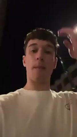 A young man is walking outside at night. He is wearing a white t-shirt, light blue denim shorts, and white shoes. He runs his hand through his hair. The background shows buildings and streetlights.