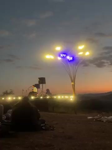 The video shows a musical performance taking place outdoors, likely on a hilltop, as indicated by the view of mountains and a city visible in the distance at dusk. A singer stands on a makeshift stage with speakers, while several people sit on the grass, facing the stage and singer. A decorative light structure resembling a tree with multiple branches and lights, stands prominently to the right of the stage. The sky is in the midst of a sunset, showing various shades of orange and blue. The mood appears relaxed and peaceful.
