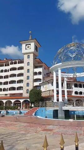 The video presents a daytime shot of the exterior of a building complex, likely a hotel or resort, under a clear blue sky with scattered clouds. The building is multi-storied, predominantly white with stone accents and reddish-brown tiled roofs. A prominent clock tower stands on the left side of the frame. In the foreground, there is a decorative gazebo-like structure with white columns and an ornate metal dome. A pool is visible in the midground, separating the viewer from the building. The camera pans upwards to focus on the clock tower and then shifts to the gazebo, providing a close-up view of its design. The focus then returns to the overall scene, showing the building, gazebo, and pool. A short, decorative fence runs along the bottom of the frame.
