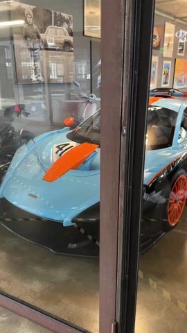 The video shows a view through a glass window into a room that appears to be a car dealership or museum, featuring several high-end sports cars. The closest car to the window is painted in a distinctive light blue with orange and black accents. The number "41" is prominently displayed on its side. Behind it, another sports car, possibly silver, can be seen. The room is decorated with artwork and posters on the walls. Lighting fixtures are visible on the ceiling.  There is writing on the glass above the cars, reading "Poccino Espresso & Service."