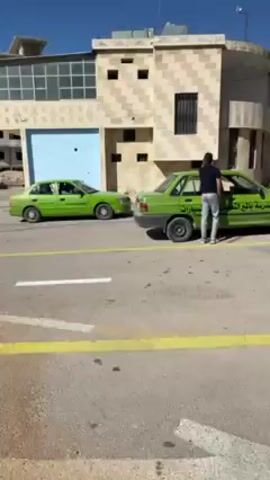 The video shows an outdoor scene with a light green car parked on a paved surface marked with white lines. There are several people standing around the car, appearing to be engaged in conversation or preparation. Another similar green car is parked nearby. In the background, there is a building made of light-colored stone and a hillside with a partially constructed building. The weather appears sunny and clear.
