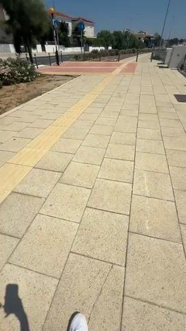 The video shows a person's point of view walking along a paved sidewalk. The sidewalk is made of light-colored square tiles and has a yellow tactile paving strip running down the middle. To the left of the sidewalk is a patch of dirt with some low-lying plants and a tree. Beyond that is a street with buildings and trees. To the right of the sidewalk is a metal railing and some modern-looking white boxes. The sky is clear and blue. The video appears to be taken on a sunny day.