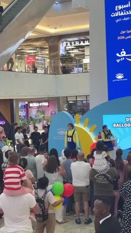 The video shows a stage set up in a shopping mall with a crowd of people watching a performance. On stage, there are two performers, one dressed in a yellow and blue outfit and the other in a blue dress. The performer in yellow appears to be interacting with the audience. The stage is decorated with a blue backdrop featuring a sun graphic and text in Arabic and English, including "DSS BALLOON". A table with some items is also visible on the stage. The crowd consists of people of various ages, some are standing, while others are holding children. In the background, there are shops and escalators visible, indicating the indoor mall environment. Balloons in red, green, and yellow are being held by someone in the foreground.