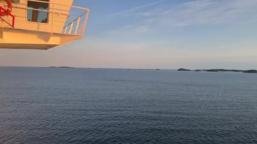 The video shows a wide expanse of calm blue water with a few small islands visible in the distance. The sun is setting on the right side of the frame, casting a warm glow across the sky and reflecting on the water. A portion of a white ship or ferry is visible in the upper left corner of the frame, suggesting the viewpoint is from a vessel. The sky is a mix of blue and soft orange and pink hues, with some wispy clouds. The overall scene is tranquil and serene.
