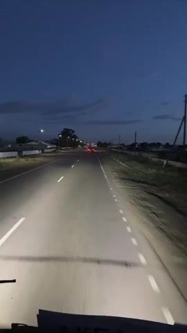 The video shows a view from inside a vehicle, likely a bus or truck, driving on a paved road at dusk. The headlights of the vehicle illuminate the road ahead, casting bright beams. White lane markings are visible on the road. On either side of the road, there is sparse vegetation and some buildings. In the distance, there are a few red lights, possibly from other vehicles. The sky is a deep blue with some clouds. In the upper portion of the video, a screen is visible, displaying a movie scene with a couple embracing. The screen appears to be mounted above the windshield of the vehicle, creating a split-screen effect with the road view. The scene suggests the occupants of the vehicle are watching a movie while traveling.
