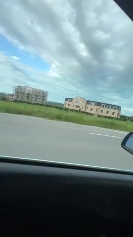 The video shows a view from inside a car driving down a road. The camera starts by looking out the passenger side window at a large, light-colored building, possibly a residential complex, set back from the road in a grassy area. The car then shifts perspective to look through the windshield, showing the road ahead. The sky is cloudy. The road is a two-lane highway with a white double line separating traffic. There are green fields on either side of the road, with trees dotted along the edges. As the car continues driving, a sign comes into view indicating the location: Krusevo/Kicevo. The dashboard of the car is visible, showing the speedometer and other gauges.
