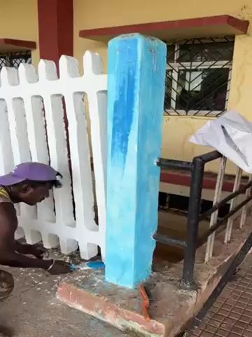 The video shows a man painting a concrete post light blue. The post is next to a white picket fence and a black metal railing. The building behind the post is yellow with maroon trim. The man is wearing a purple hat and is using a paintbrush to apply the paint to the post. There is an orange tool on the ground near the base of the post, and a white plastic bag is draped over the metal railing. The ground is tiled.
