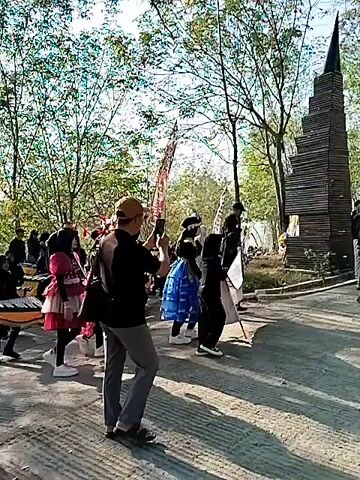 The video shows a group of people, including children, walking outdoors in what appears to be a parade or procession. The children in the foreground are dressed in black shirts and pants with traditional patterned sashes, and they are carrying and playing drums. There are also flags with writing on them displayed. In the background, there are more people, trees, and a tall, dark, abstract sculpture made of stacked materials. The ground is paved and there are some plants and foliage visible. The overall atmosphere seems festive.