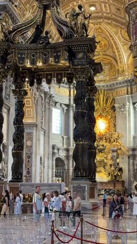 The video displays the interior of a grand basilica, centered on Bernini's Baldachin. The Baldachin is a large, dark bronze and gold structure with four twisted columns supporting an ornate canopy adorned with statues. In the background, Bernini's Altar of the Chair is visible, a gilded edifice featuring a central stained-glass dove surrounded by golden rays and flanked by statues. The basilica's high, intricately decorated golden dome is visible overhead. Numerous tourists, casually dressed, are scattered around the Baldachin, standing and observing the architectural features. Some individuals are in the immediate foreground, positioned behind a clear barrier. Natural light illuminates the scene, entering through a tall arched window located to the left of the Baldachin.