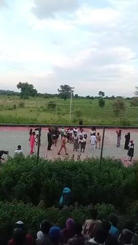 The video shows a group of people on a tennis court, some dancing and others observing. The court is surrounded by a fence and a lush green landscape with trees in the background. The sky is overcast. In the foreground, a crowd of people is gathered, watching the activity on the court.