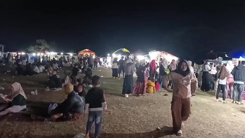 The video depicts a lively nighttime outdoor gathering with numerous people scattered across a dry, grassy field. In the background, a row of stalls or tents illuminated by bright lights creates a vibrant marketplace atmosphere. People are seen browsing at the stalls and socializing. In the foreground and midground, individuals and small groups are seated on the ground, some appearing to be eating or conversing. Children are also present, with one child walking in the center of the frame. The overall scene suggests a festive community event or market taking place after dark.