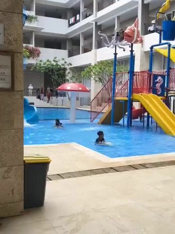 The video shows a children's swimming pool area with a water slide and playground features. Two children are visible in the pool. One child is swimming, and the other is playing with a yellow toy in the shallow end. A red mushroom-shaped water feature is in the middle of the pool, and a water bucket feature with a fish decoration is on the right side of the slide. The pool area is surrounded by a building with balconies, and there are some trees and plants visible. A sign with pool water quality information is mounted on a pillar to the left of the frame. A trash bin is also located next to the pillar.